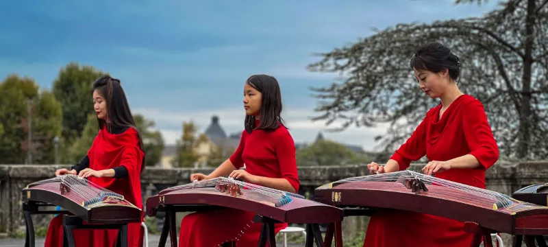 Three guzheng players in red dresses performing side by side outdoors